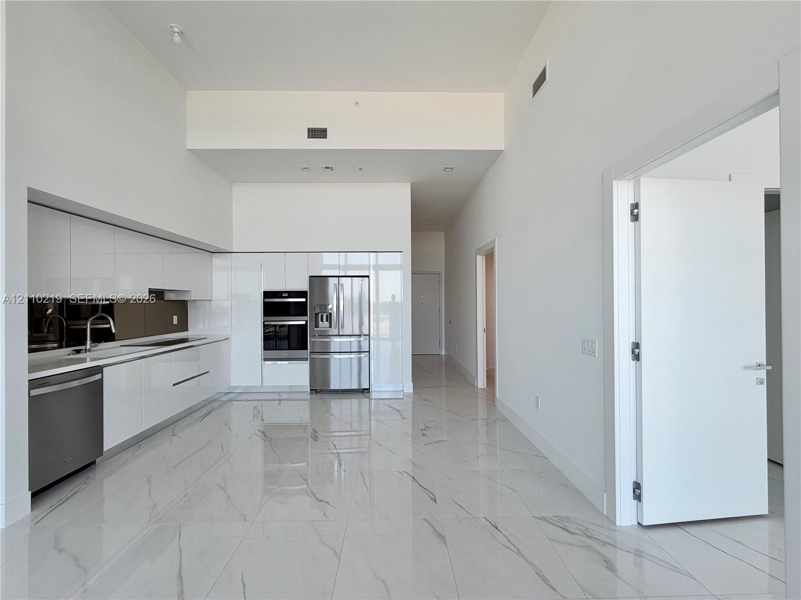 Contemporary kitchen featuring high-gloss white cabinetry, integrated stainless steel appliances, polished stone-finish tile flooring, and soaring ceilings
