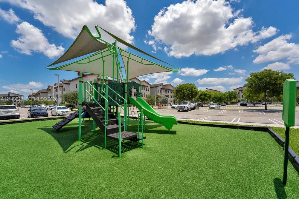 Playground featuring a shade structure, metal slide, and climbing equipment situated on a synthetic turf surface with a defined border