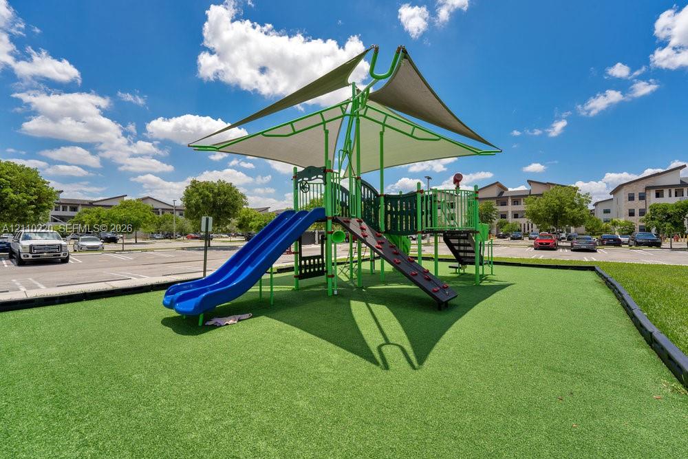 Outdoor recreational structure featuring dual slides, a climbing wall, and a tension-fabric shade canopy situated on a synthetic turf surface