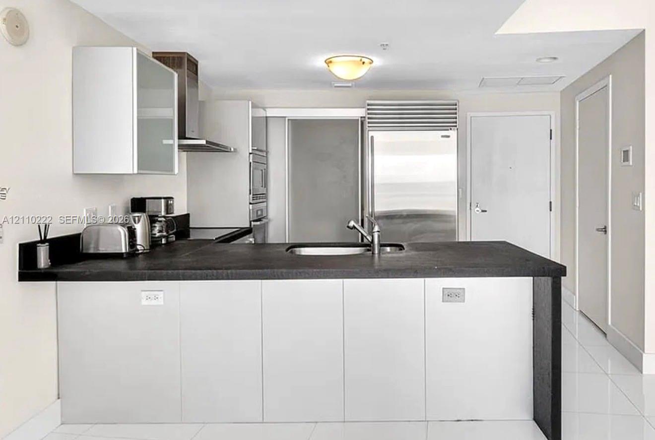Kitchen featuring flat-panel cabinetry, a stainless steel refrigerator, stone-surfaced island with an integrated sink, and stainless steel range hood Kitchen featuring flat-panel cabinetry, a stainless steel refrigerator, stone-surfaced island with an integrated sink, and stainless steel range hood