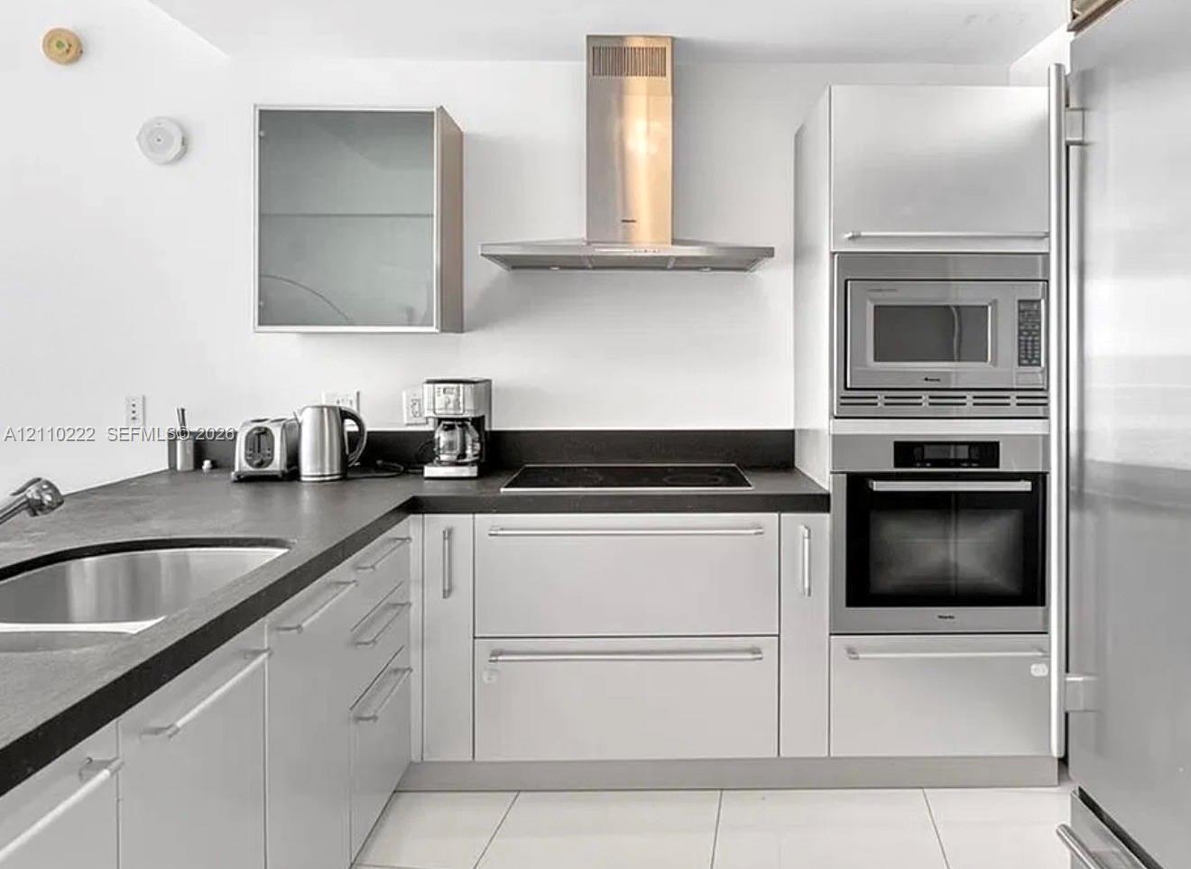 Kitchen featuring sleek white cabinetry, matte black countertops, stainless steel range hood, built-in wall oven and microwave, and large-format tile flooring Kitchen featuring sleek white cabinetry, matte black countertops, stainless steel range hood, built-in wall oven and microwave, and large-format tile flooring