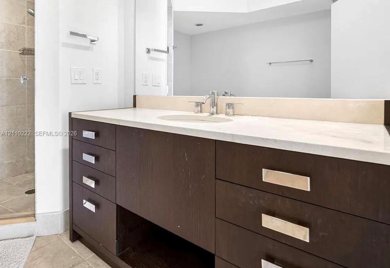 Bathroom vanity featuring dark wood-finish cabinetry, a neutral solid-surface countertop with an integrated oval sink, polished metal hardware, and a large-format mirror Bathroom vanity featuring dark wood-finish cabinetry, a neutral solid-surface countertop with an integrated oval sink, polished metal hardware, and a large-format mirror