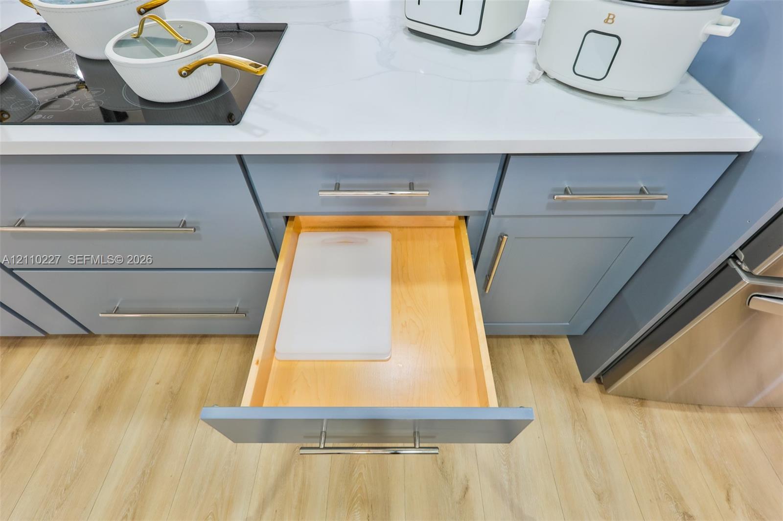 Shaker-style cabinetry with matte finish, stone countertop, built-in induction cooktop, wood-finish flooring, and cabinetry featuring pull-out drawer storage with metal hardware