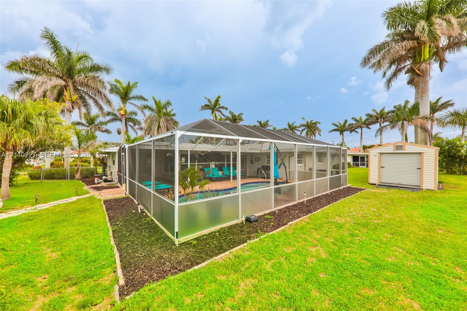 Screen-enclosed swimming pool featuring a paved deck and a detached storage shed situated on a lawn with mature palm trees
