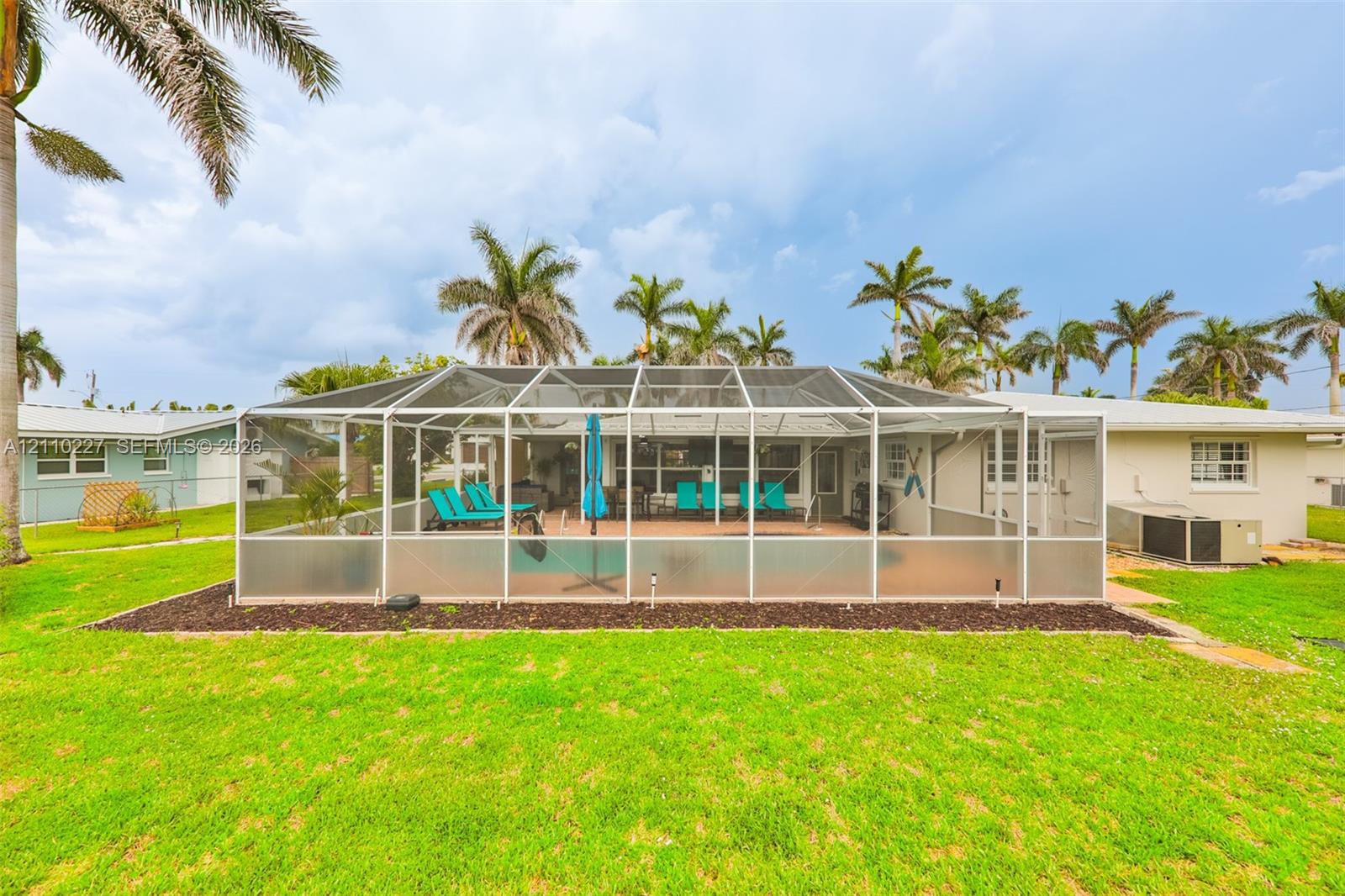Screen-enclosed swimming pool featuring a paved deck, surrounded by a manicured lawn with mature palm trees