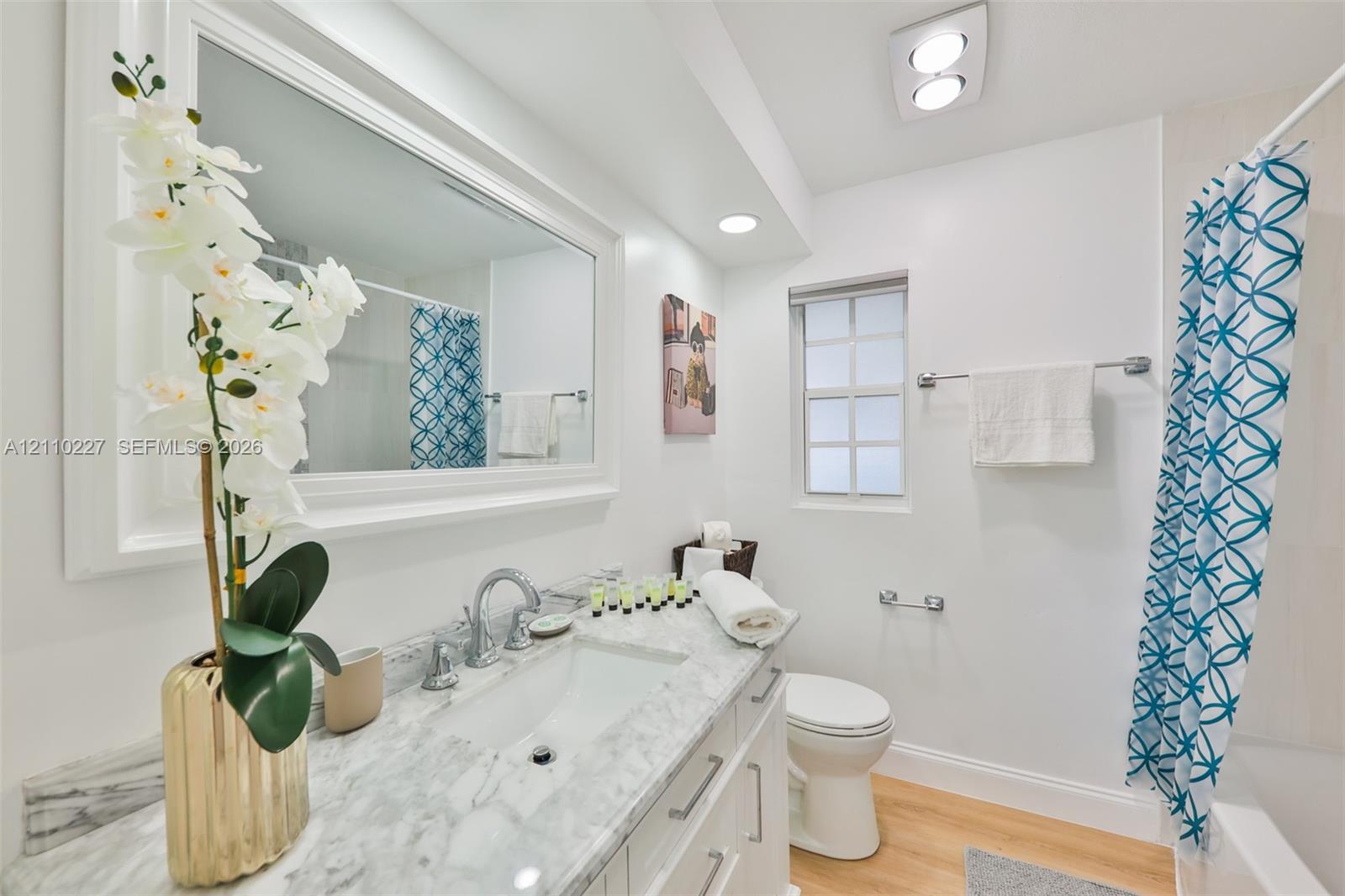 Bathroom featuring a stone-top vanity with an undermount sink, framed wall-to-wall mirror, wood-finish flooring, recessed ceiling lighting, and a multi-pane window