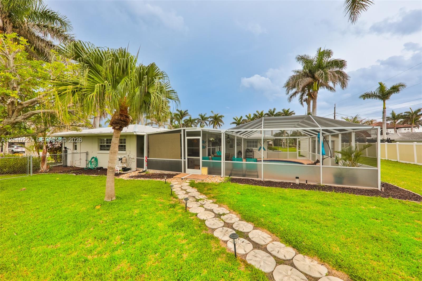 Expansive backyard featuring a manicured lawn, stone paver pathway, mature palm trees, and an enclosed swimming pool with a screened lanai