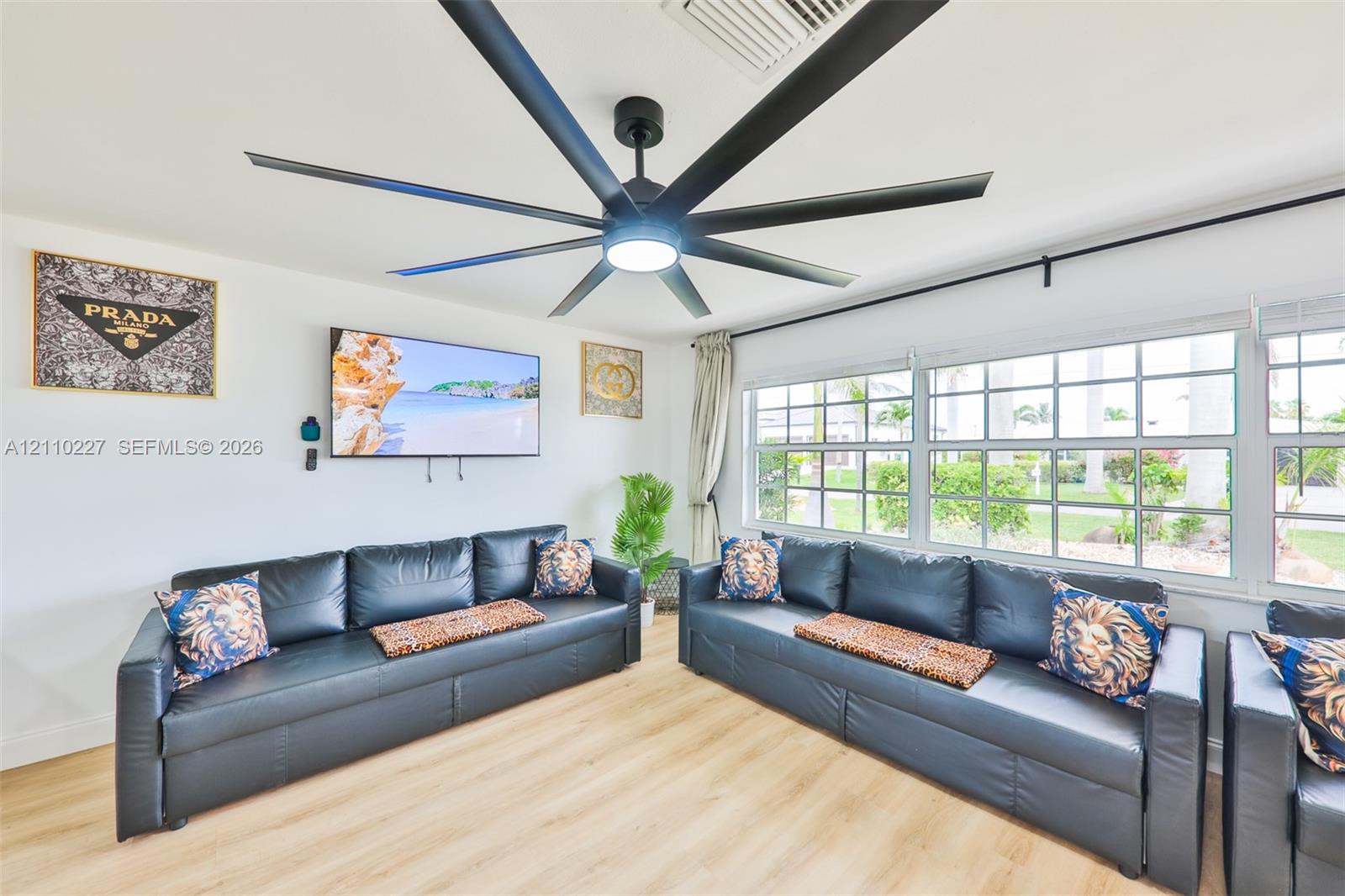 Multi-blade matte black ceiling fan with integrated lighting, wood-finish flooring, and expansive grid-pane windows
