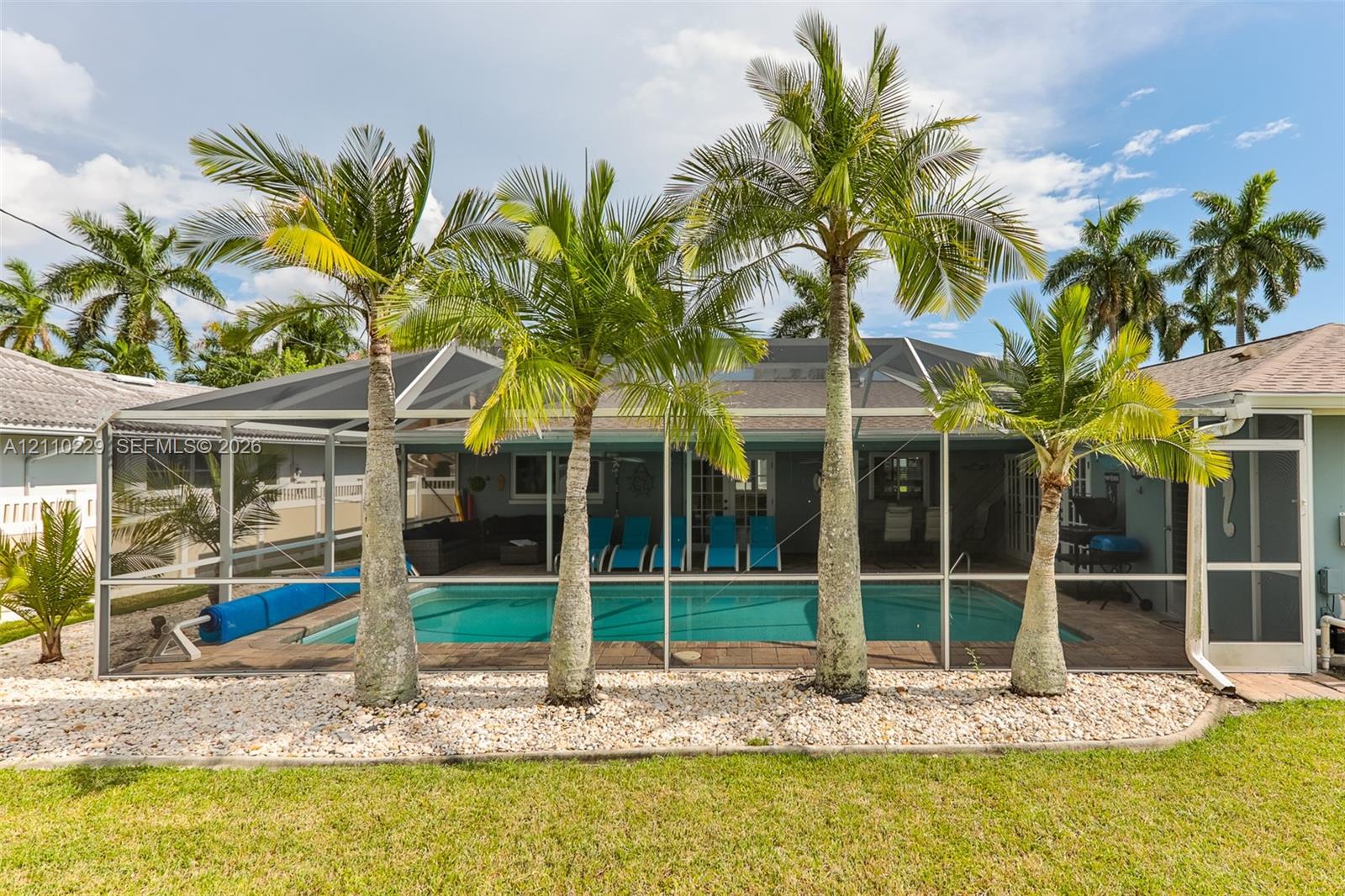 Screened-in outdoor swimming pool featuring paver decking, mature palm trees, and light stone ground cover