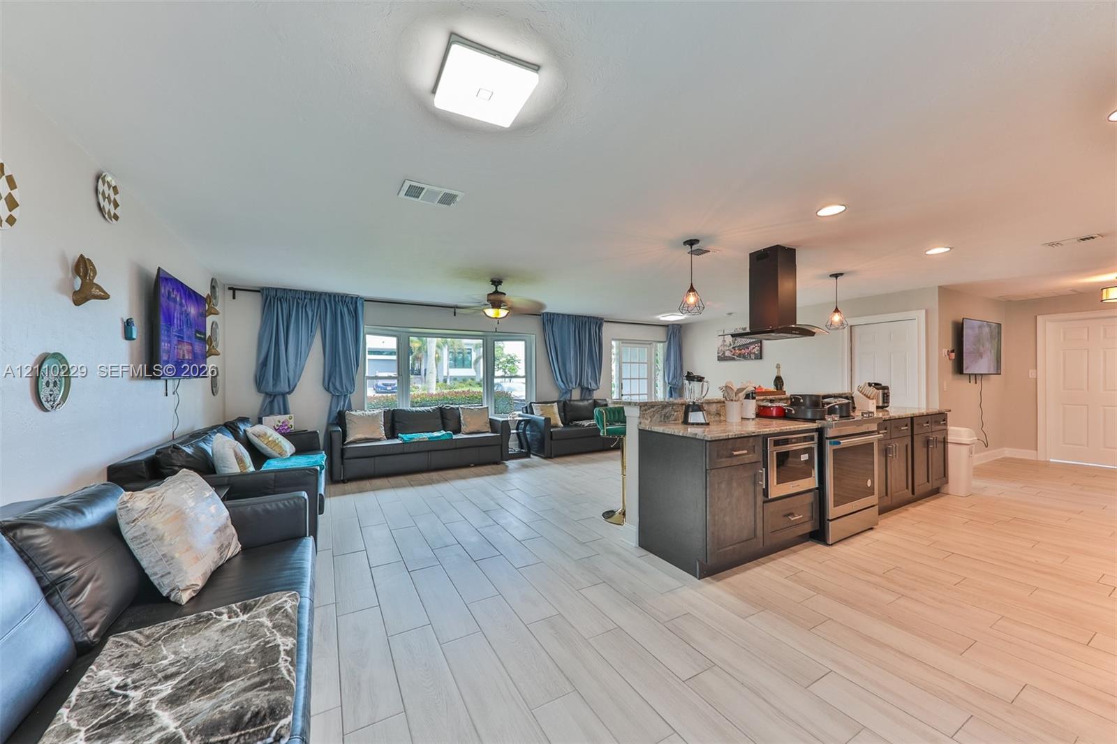 Open-concept interior featuring light-toned tile flooring, dark-finish cabinetry, stone-style countertops, stainless steel kitchen appliances, and a modern ceiling-mounted range hood