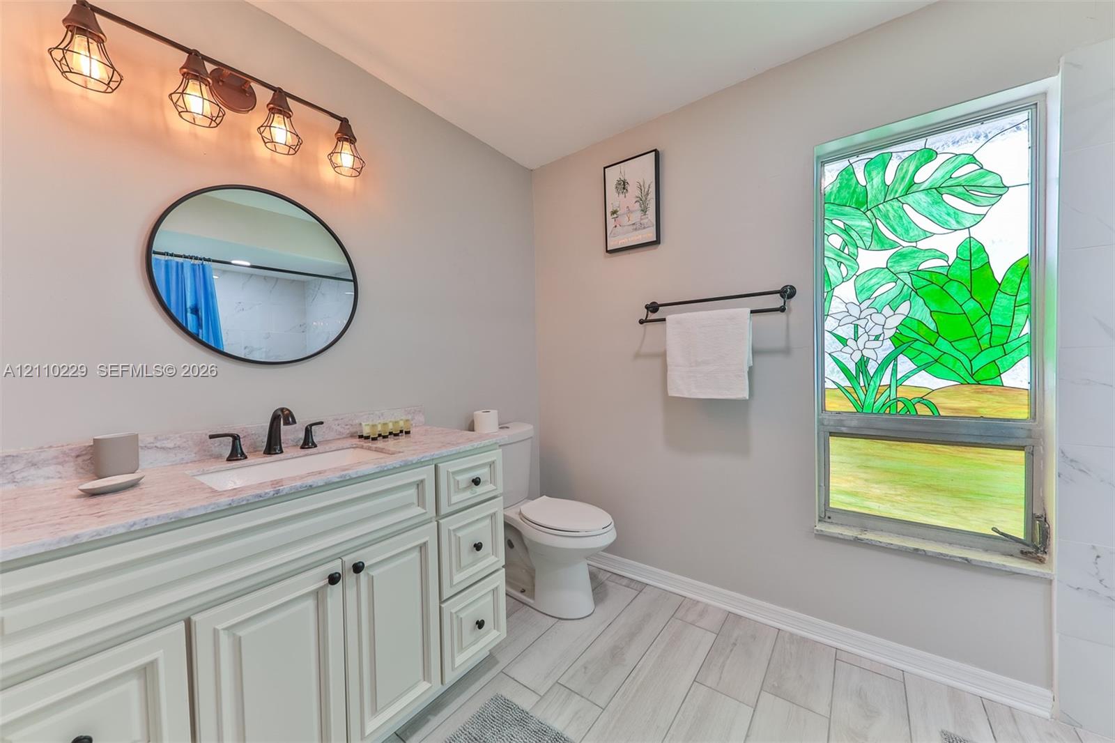 Bathroom featuring a custom stained-glass window, vanity with stone countertop, oval wall-mounted mirror, industrial-style four-light fixture, and wood-finish flooring