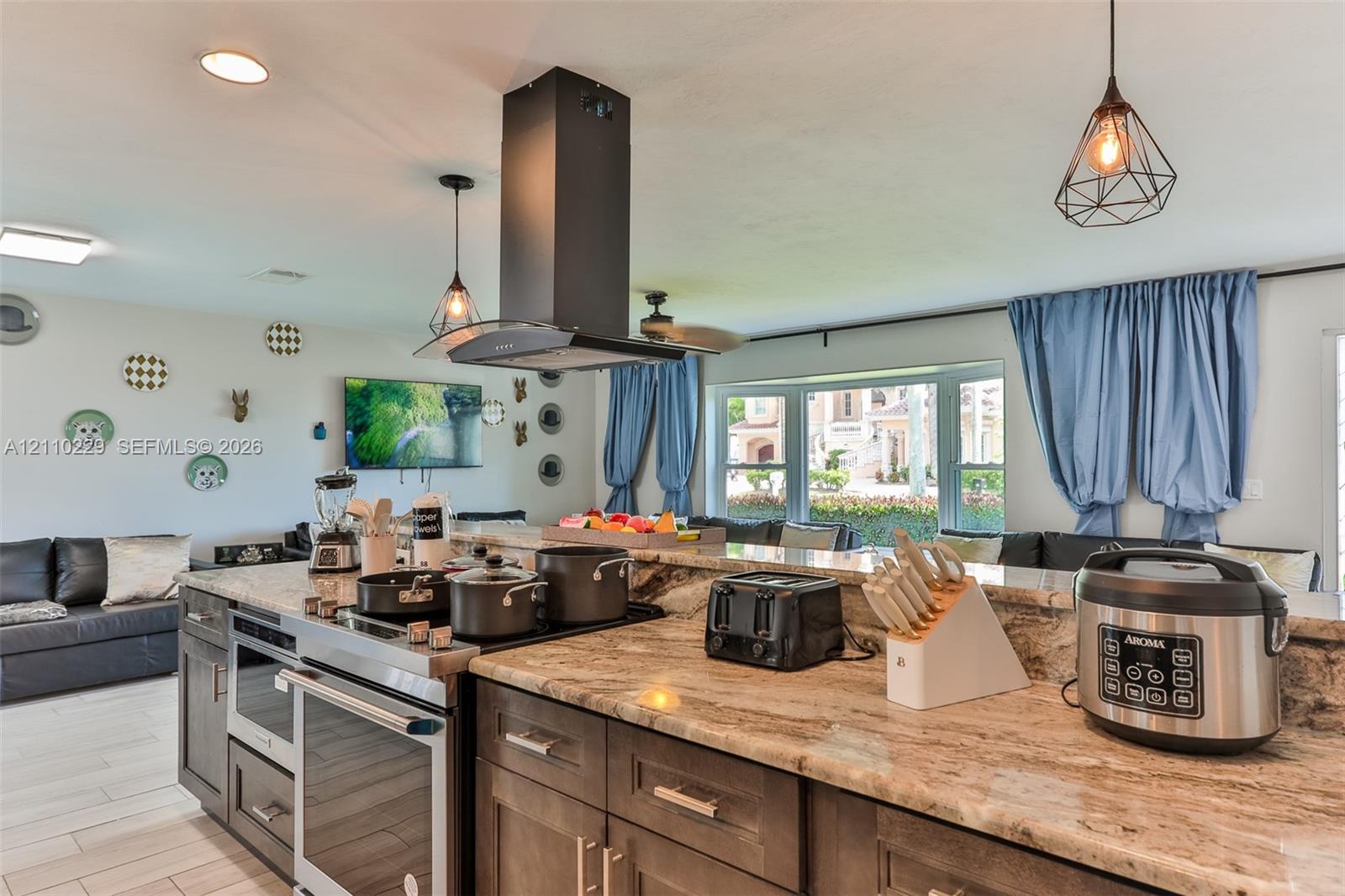 Kitchen featuring stone countertops, dark-finish cabinetry, an integrated cooktop with a stainless steel chimney-style range hood, tile flooring, and geometric pendant lighting