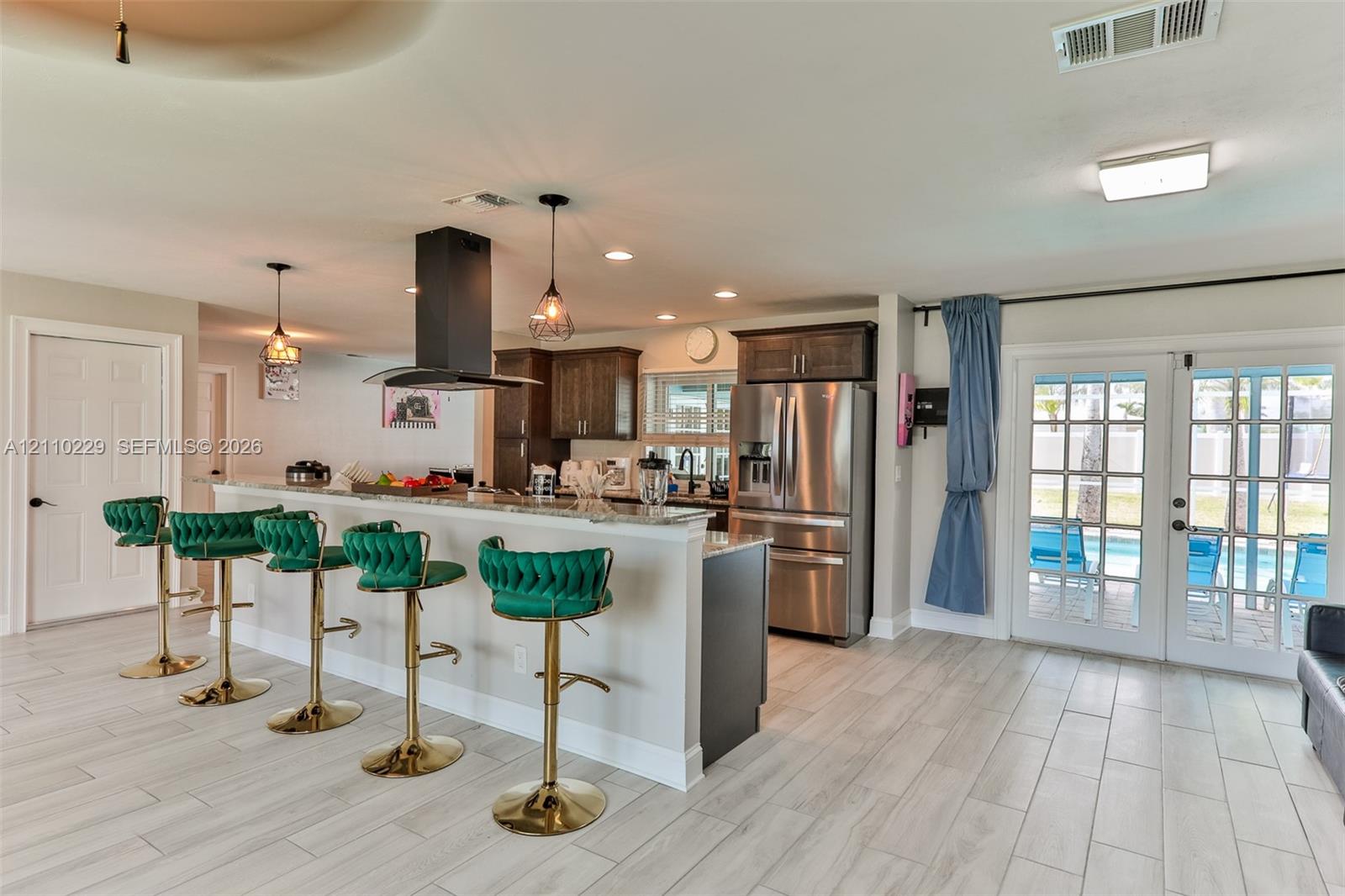 Kitchen featuring wood-finish cabinetry, stone-surface breakfast bar, stainless steel refrigerator, matte black chimney-style range hood, and double glass-paned doors
