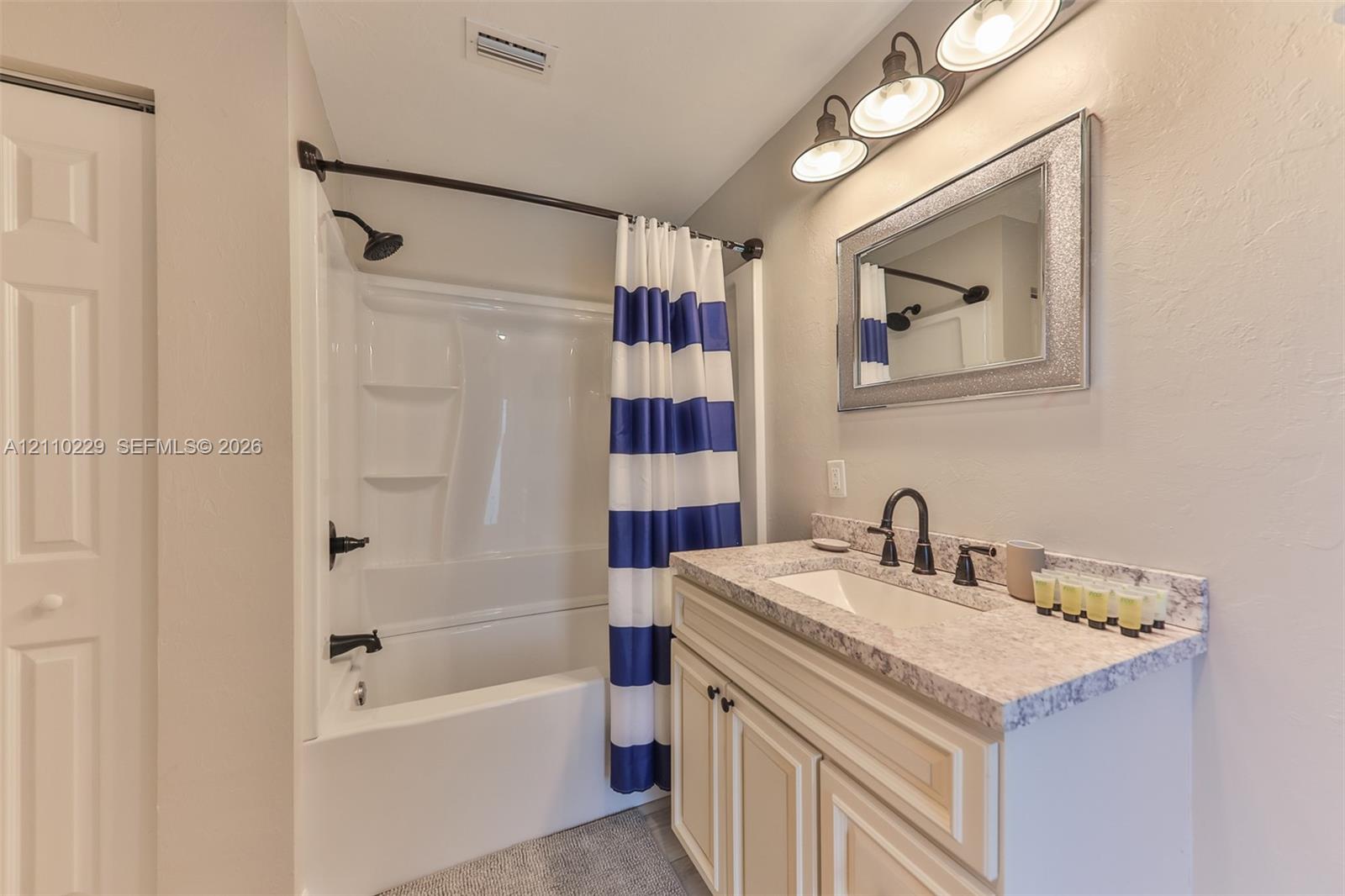 Vanity featuring a stone-composite countertop, integrated rectangular sink, oil-rubbed bronze faucet, and shaker-style cabinetry, complemented by a tub-shower combination with matching bronze fixtures and a three-light vanity fixture