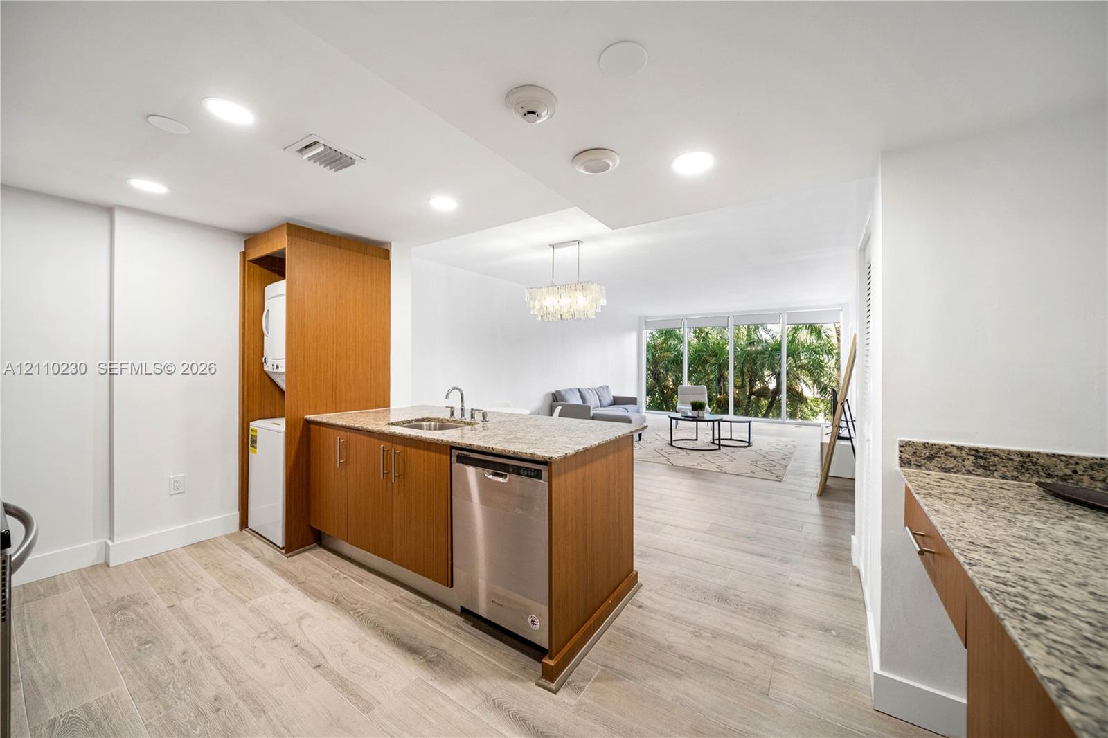 Kitchen featuring stone-finish countertops, stainless steel dishwasher, wood-finish cabinetry, wood-finish flooring, and a built-in laundry station with stacked unit