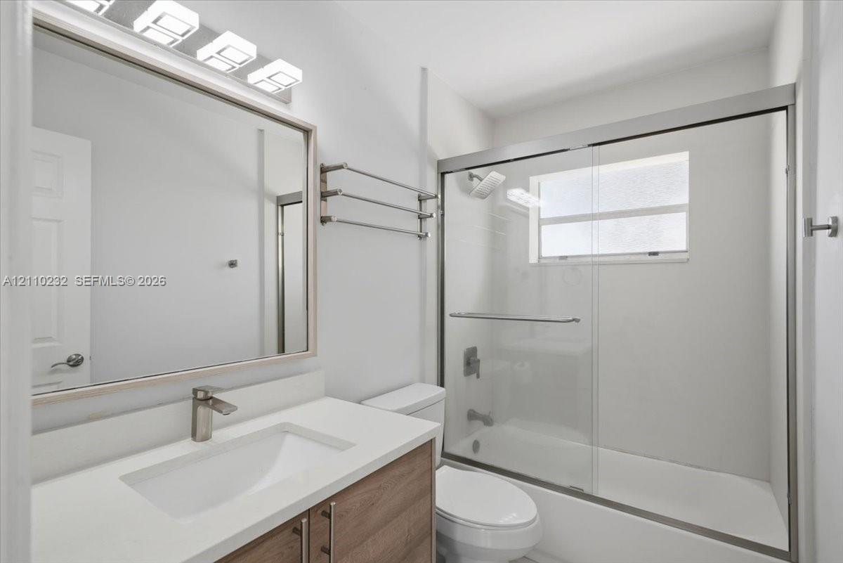 Wood-finish vanity with white countertop, integrated sink, brushed-metal hardware, sliding glass shower enclosure, and three-light wall-mounted fixture