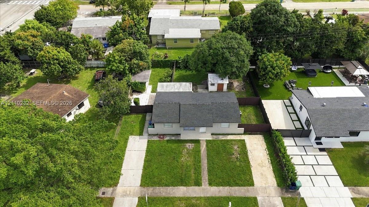 Aerial perspective featuring a detached residential structure with a shingled roof, a paved concrete driveway, and a fenced yard with mature trees