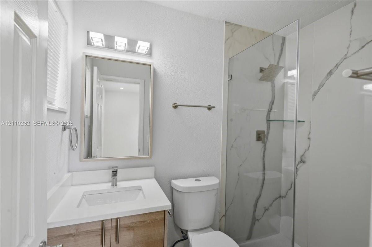 Bathroom featuring a wood-finish vanity with a rectangular sink, quartz countertop, wall-mounted mirror, frameless glass shower enclosure, and white tile with dark veining