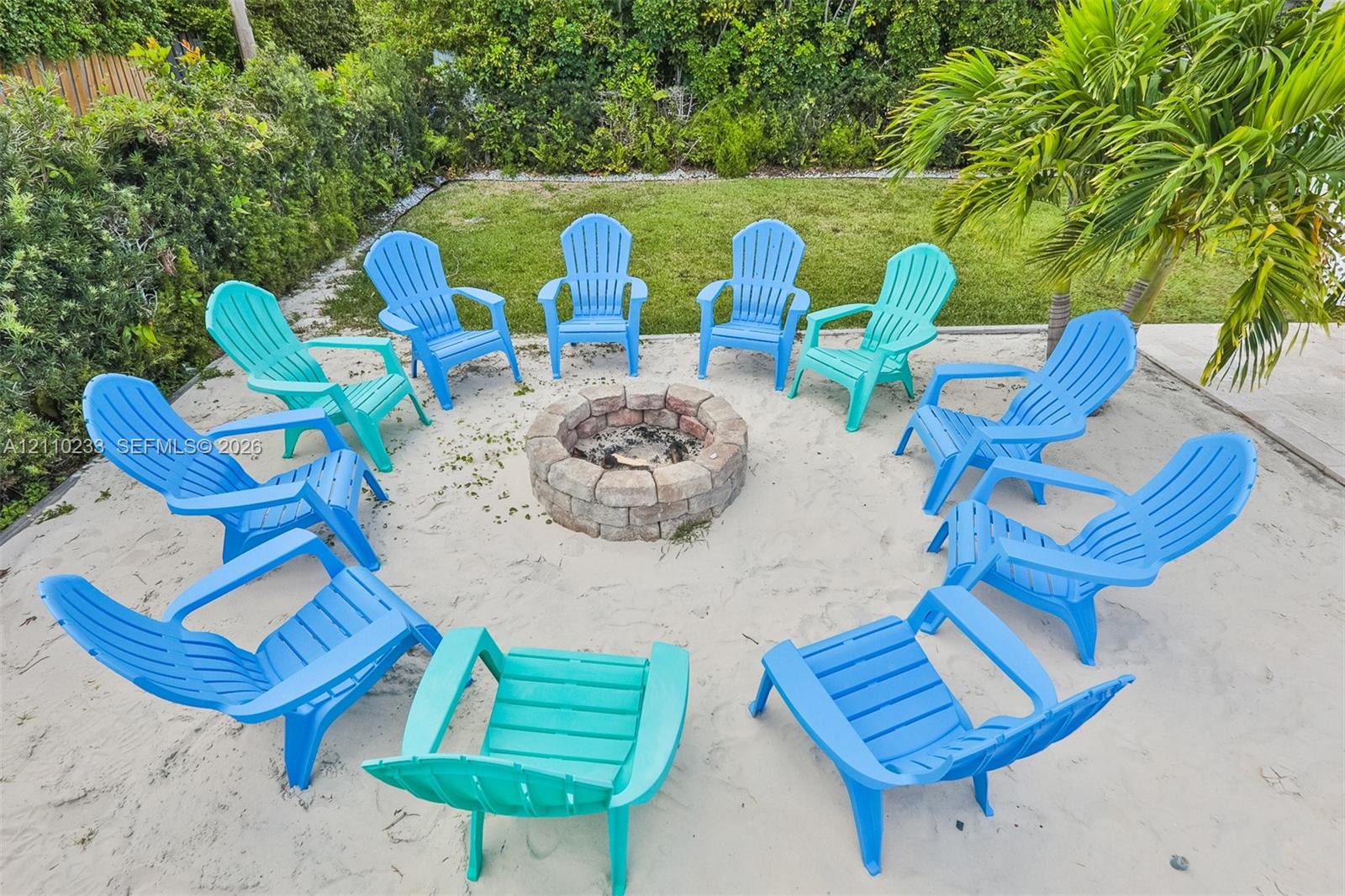 Backyard featuring a circular stone fire pit, sand ground cover, manicured lawn, and perimeter hedging