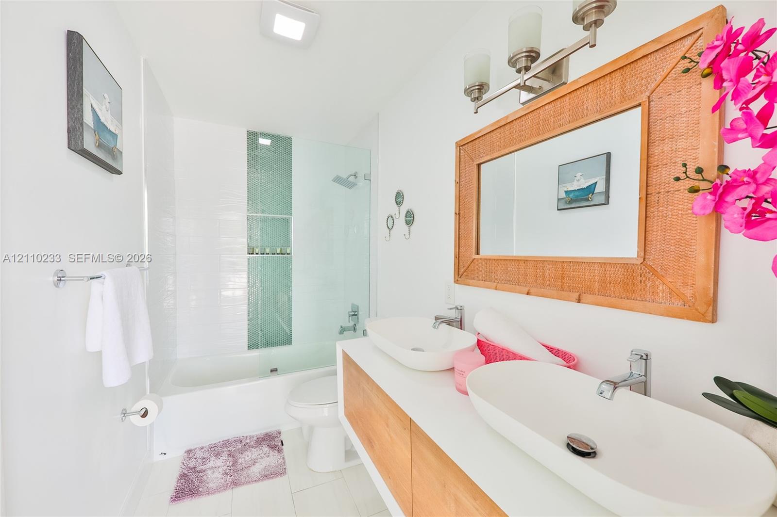 Floating double vanity featuring light wood-finish cabinetry, rectangular vessel sinks, and polished chrome hardware, complemented by a glass-enclosed shower with a vertical mosaic tile accent