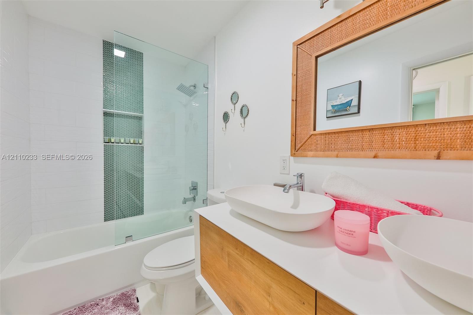 Bathroom featuring a wood-finish floating vanity with dual vessel sinks, a glass-enclosed tub shower with mosaic tile inlay, and a built-in wall mirror