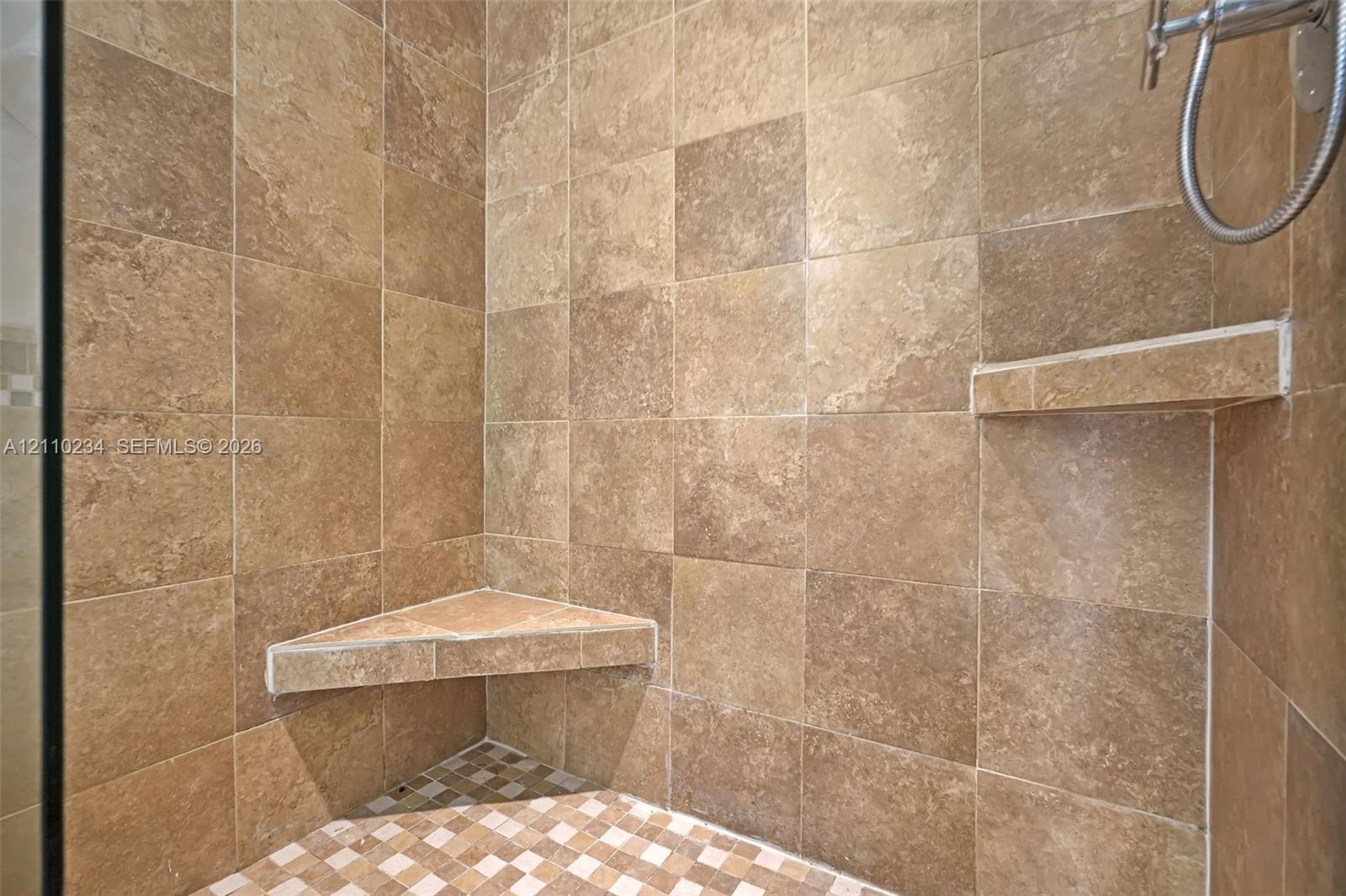 Walk-in shower featuring large-format neutral-toned tile walls, a built-in corner bench, a floating corner shelf, and a mosaic-tiled floor with metal shower fixture