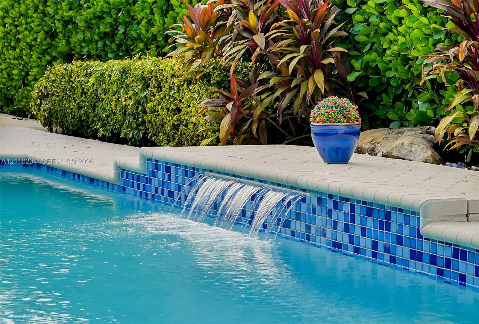 In-ground pool featuring a tiled water feature, blue glass mosaic waterline tile, light-toned concrete coping, and manicured perimeter landscaping