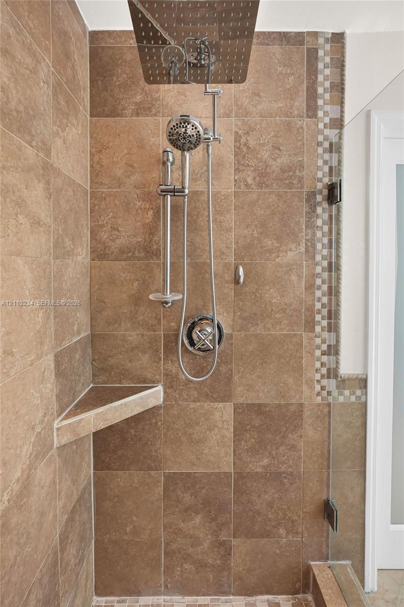 Walk-in shower featuring large-format earth-tone tile, a square rainfall showerhead with a secondary handheld sprayer, a built-in triangular corner shelf, and a vertical mosaic accent border