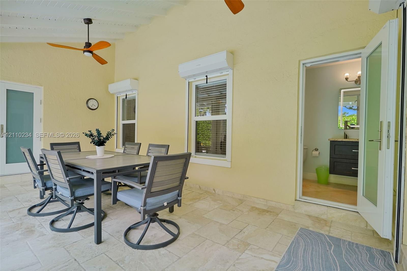 Covered patio featuring stone tile flooring, two ceiling-mounted fans, multiple glass-paneled entry doors, and wall-mounted ductless climate control units