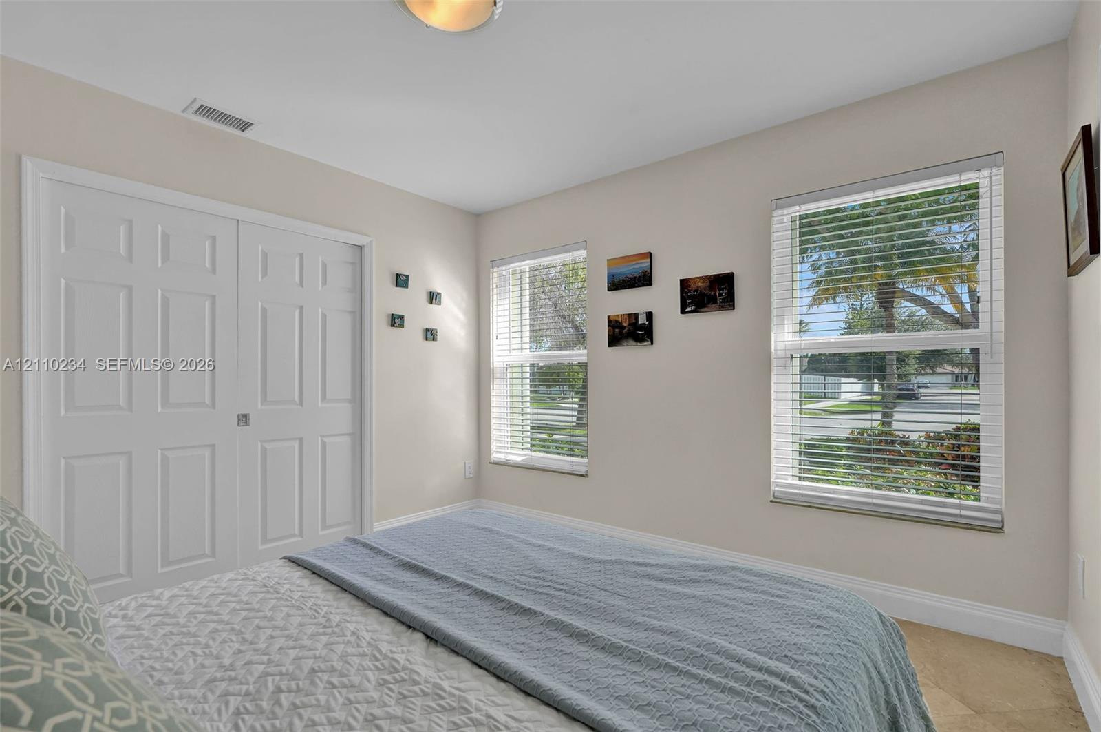 Neutral-toned walls, double-door closet with recessed paneling, and dual windows equipped with horizontal blinds, complemented by light-colored tile flooring and a flush-mount ceiling light fixture