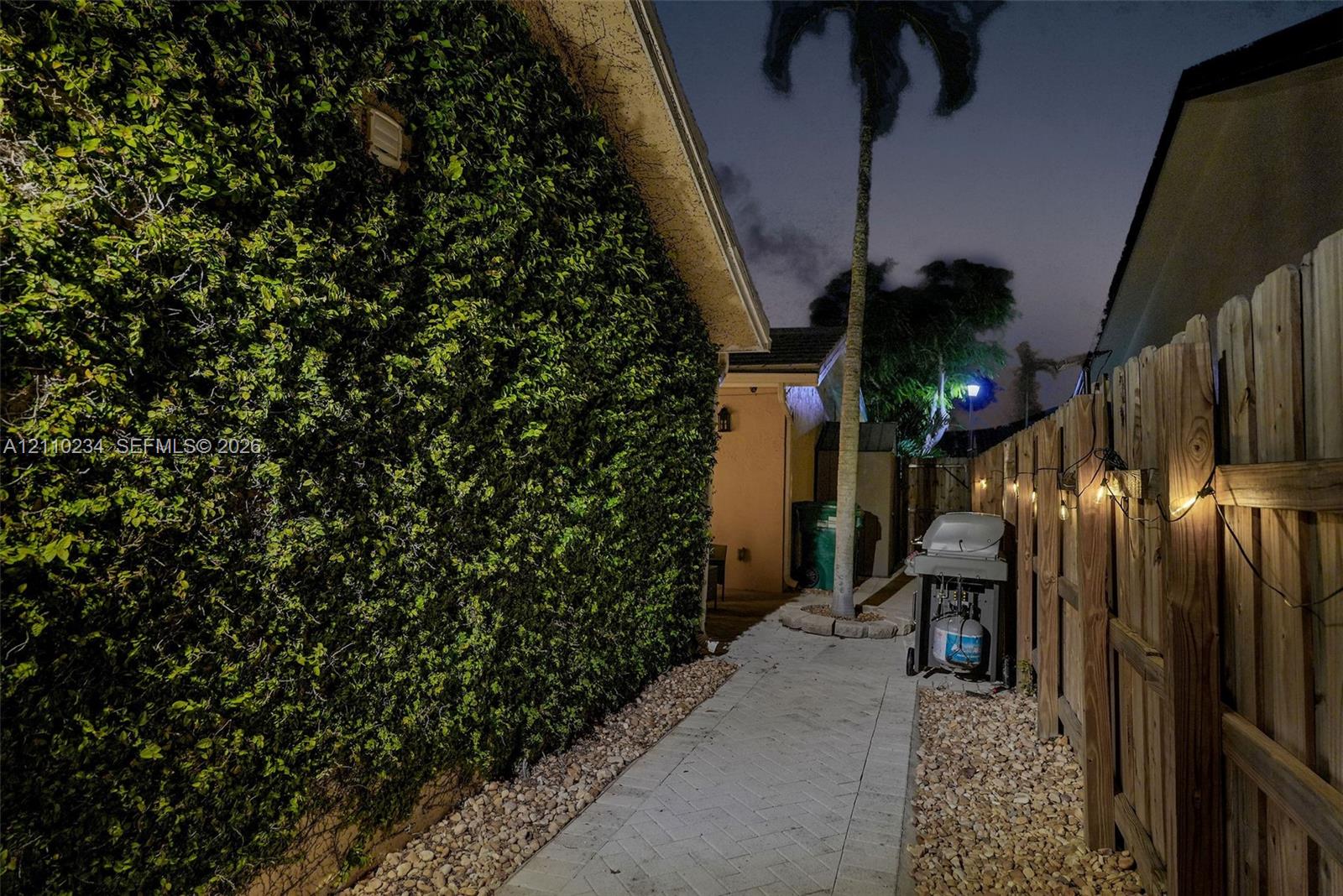 Exterior side walkway featuring a paved stone path, wood privacy fencing, a manicured hedge wall, and mature palm landscaping