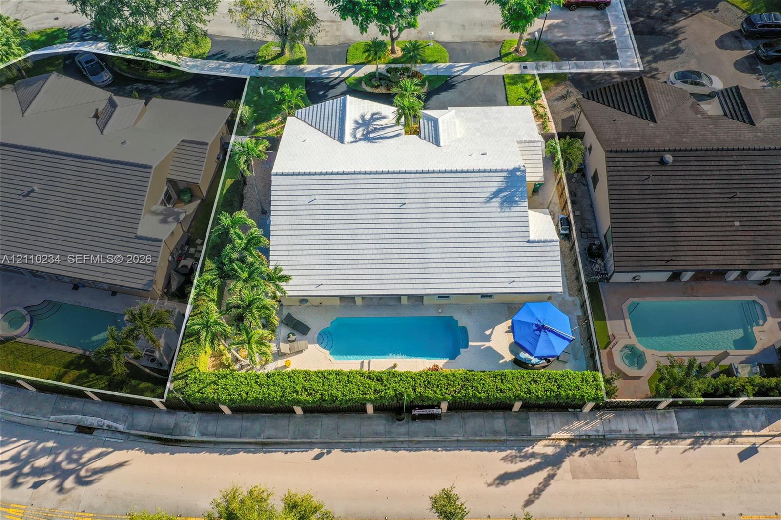 Aerial perspective of property featuring a light-toned tile roof, an in-ground swimming pool with a hardscaped deck, and mature palms within a fenced perimeter