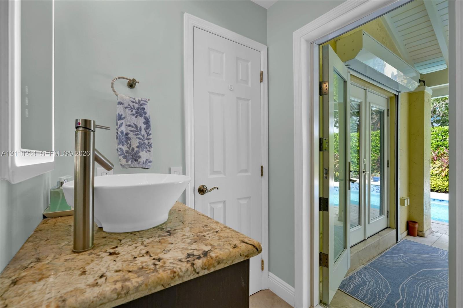 Vanity featuring a stone countertop, vessel sink with a high-arch faucet, and an integrated LED-backlit mirror, with double French doors opening to an exterior pool area