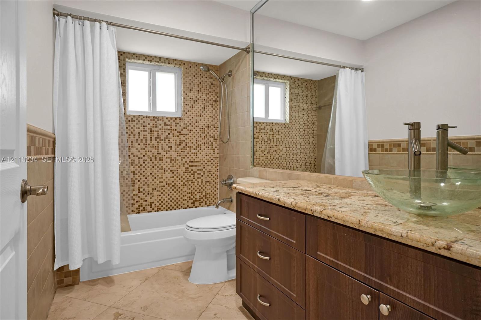 Bathroom featuring a wood-finish vanity with a stone countertop, glass vessel sink, mosaic tile tub surround, and large wall-mounted mirror