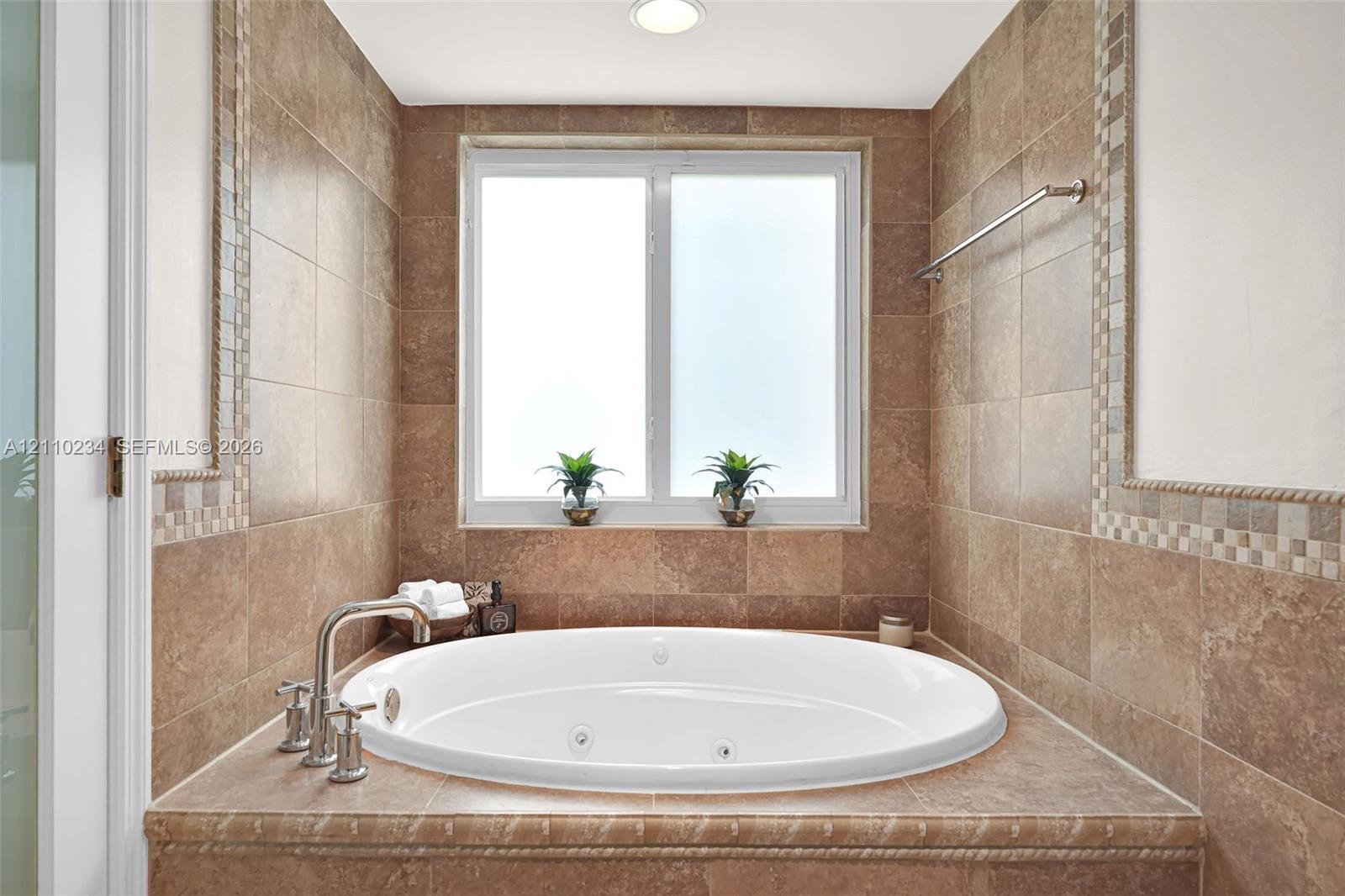 Oversized jetted soaking tub featuring a tile-surround deck, mosaic tile accents, polished chrome fixtures, and a dual-pane window