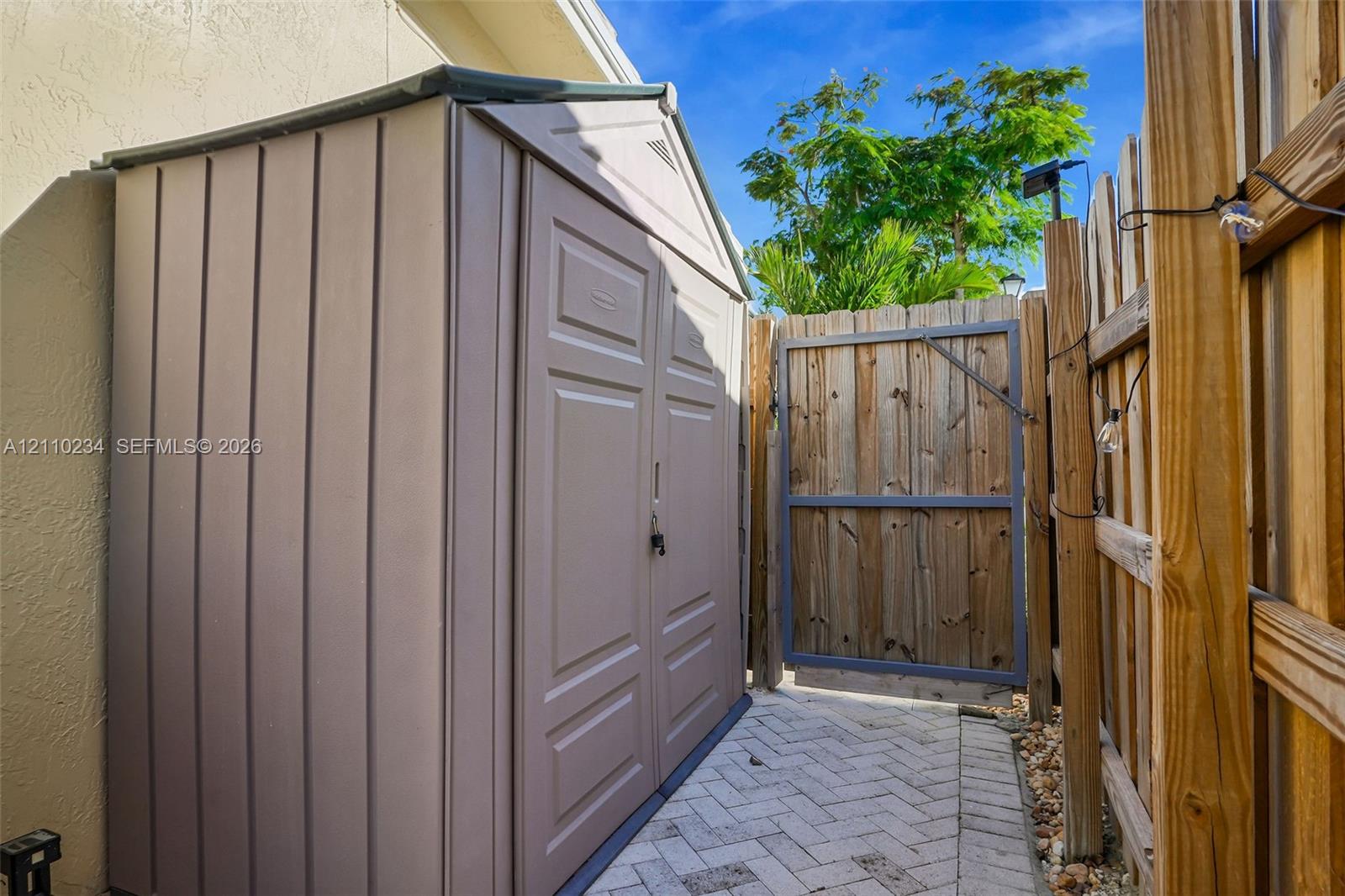 Storage shed with double doors, herringbone-patterned paver walkway, and wooden privacy fencing