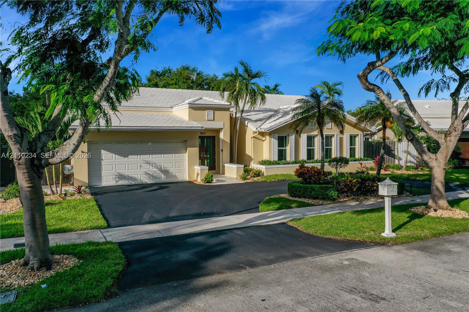 Single-story residence featuring a multi-gabled light-toned roof, an attached two-car garage, manicured landscaping with mature palm trees, and a paved driveway