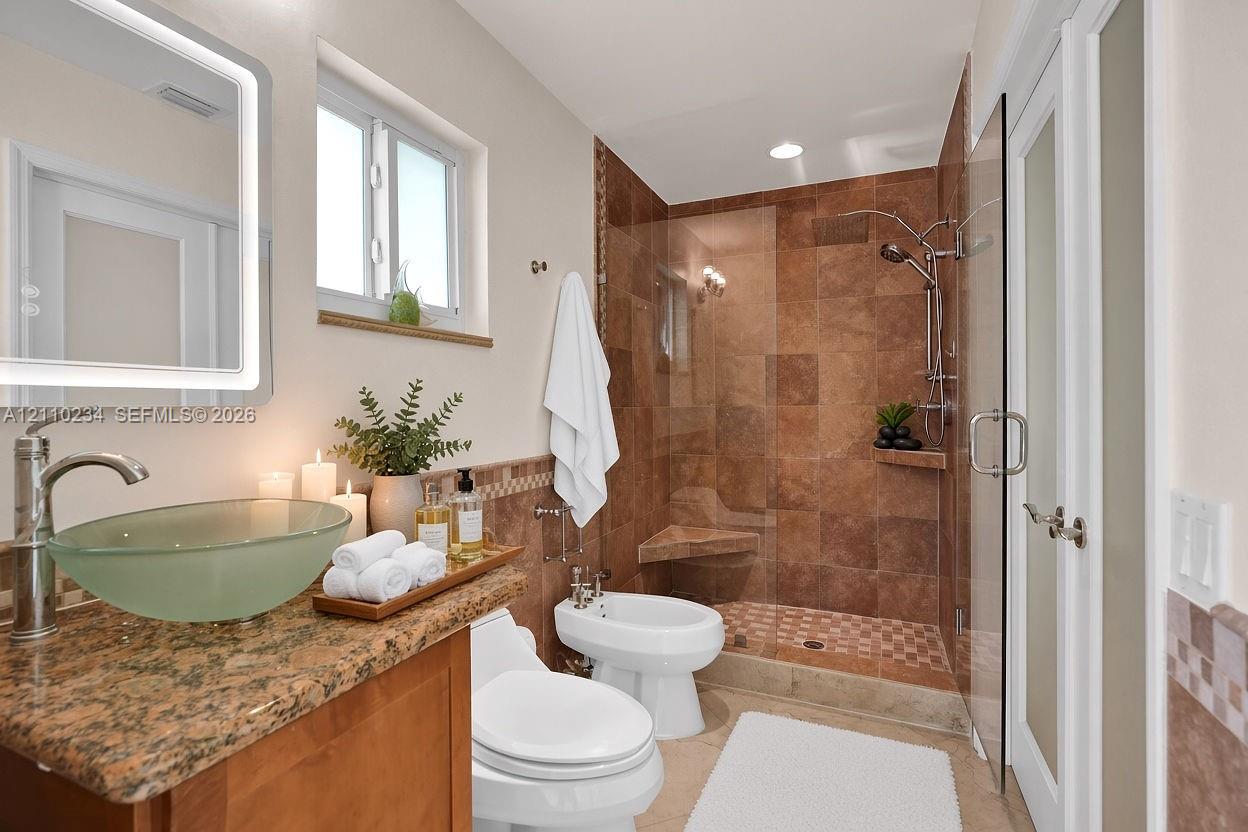 Bathroom featuring a frosted glass vessel sink, stone vanity countertop, lighted vanity mirror, and a walk-in shower with tiled walls, built-in bench, and a glass door