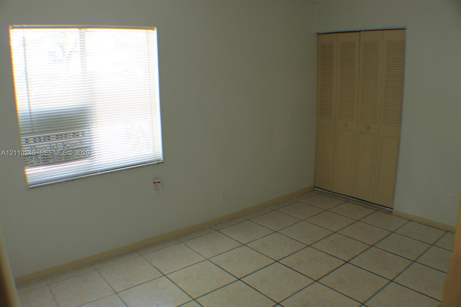 Neutral-toned interior space featuring light-colored tile flooring, window with horizontal blinds, and closet with bi-fold louvre-style doors