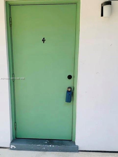 Exterior entrance featuring a solid-core door with a painted sage finish, a matte black door number, and a wall-mounted light fixture