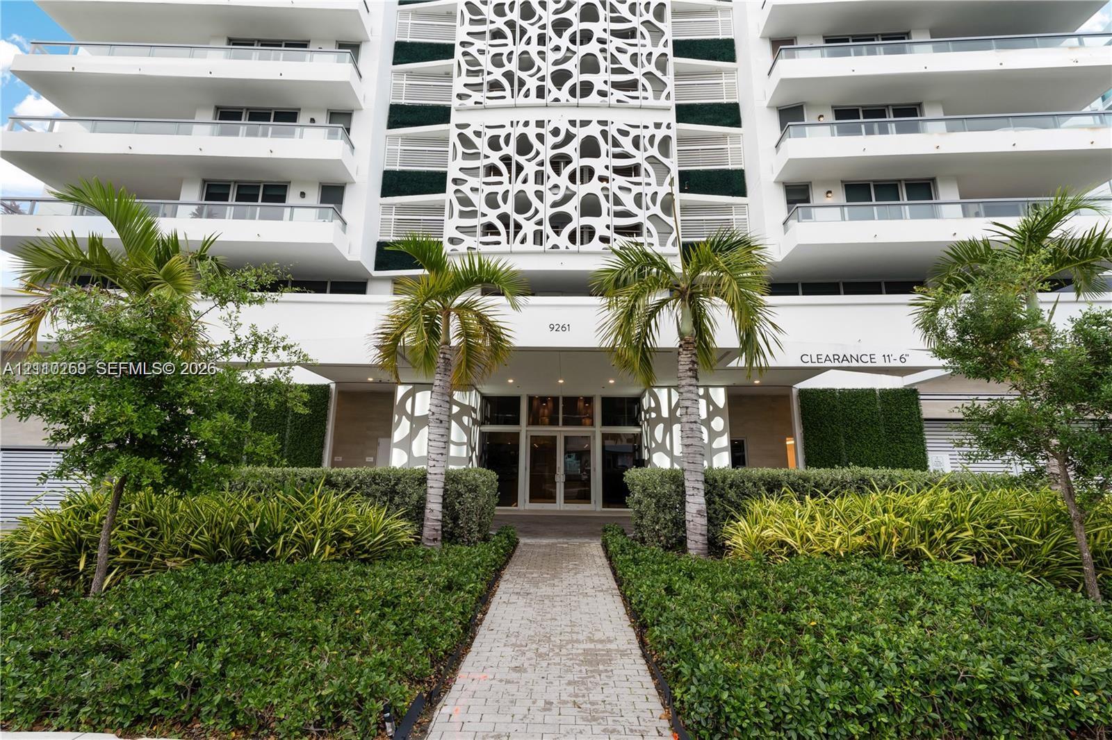 Modern mid-rise exterior featuring decorative latticework panels, tiered balconies with glass railings, landscaped hedging, and a paved stone entry path
