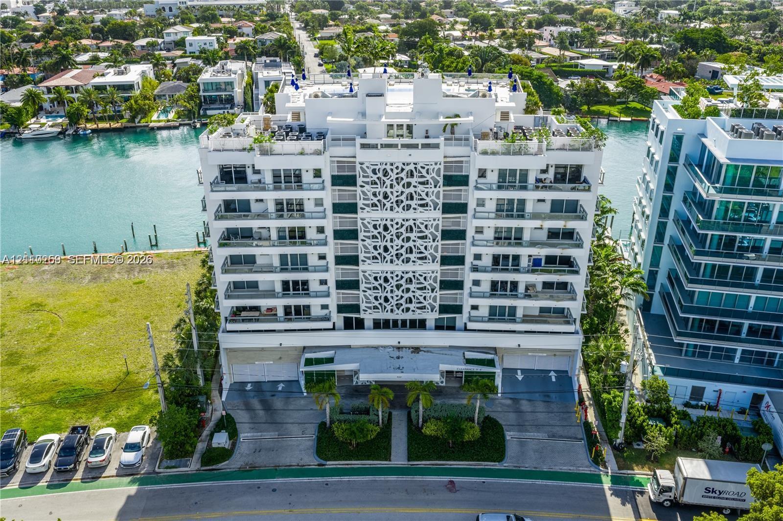 Waterfront high-rise building featuring decorative facade screening, private balcony levels, ground-level covered parking, and palm tree landscaping