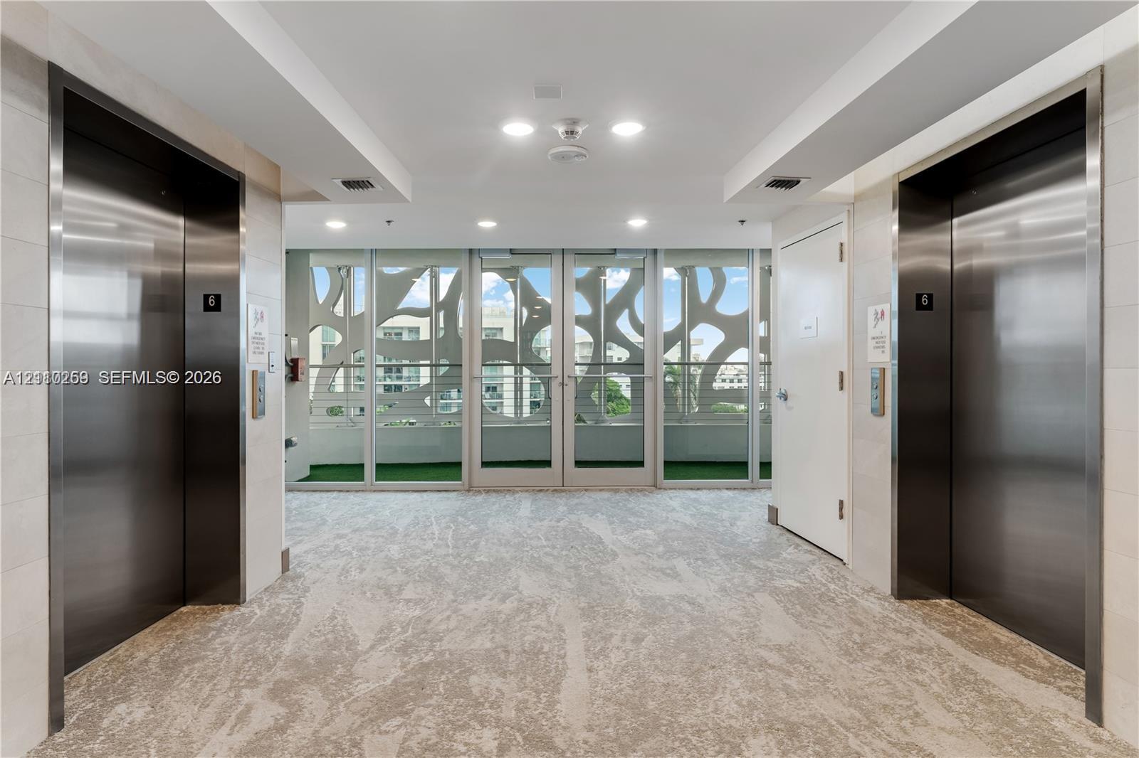 Elevator lobby featuring stainless steel doors, neutral patterned carpeting, recessed lighting, and floor-to-ceiling glass paneling with decorative metal screening
