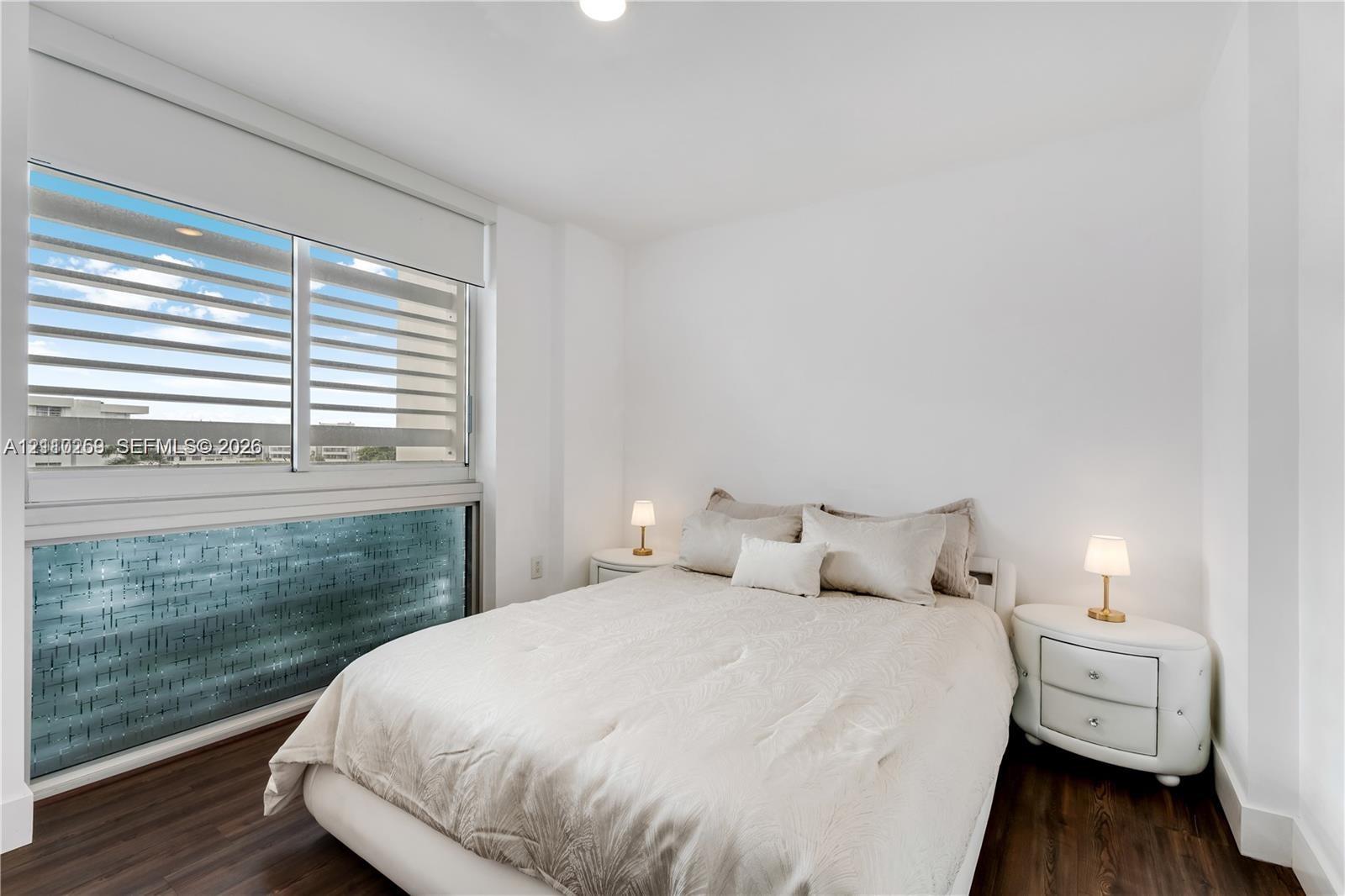 Room featuring dark wood-finish flooring, white walls, and a large horizontal window with integrated blinds
