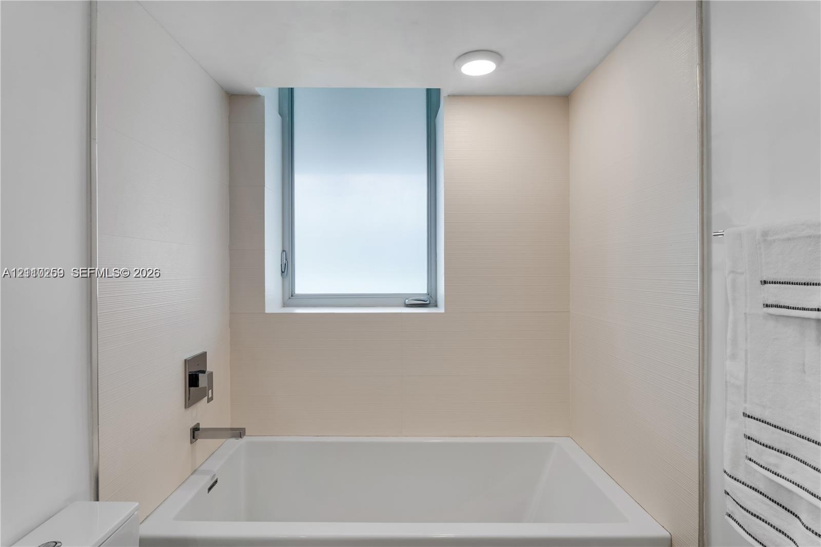 Bathtub enclosure featuring matte-finish beige wall tile, a recessed window, a rectangular-profile wall-mounted faucet, and a circular ceiling-mounted light fixture