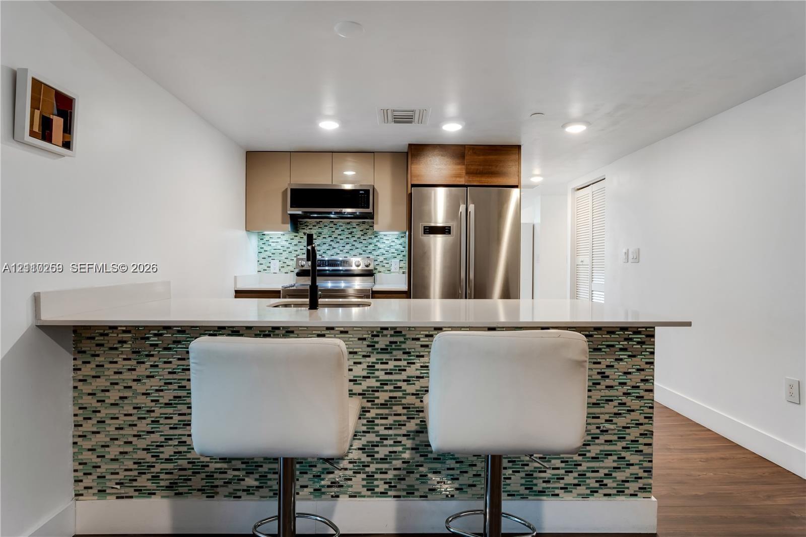 Kitchen featuring high-gloss cabinetry, stainless steel appliances, a multi-tone glass tile backsplash, and a white countertop with an integrated breakfast bar