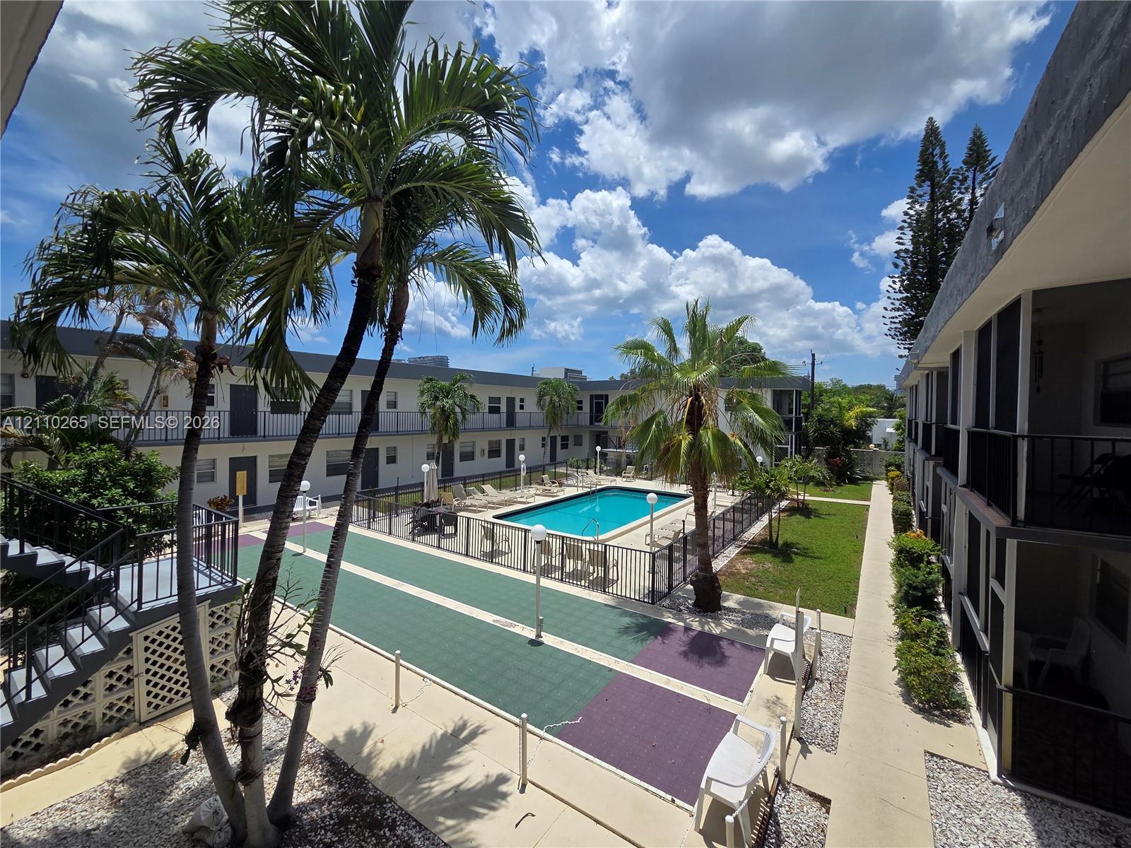 Swimming pool with security fencing, palm tree landscaping, paved recreation area, and multi-story building exterior