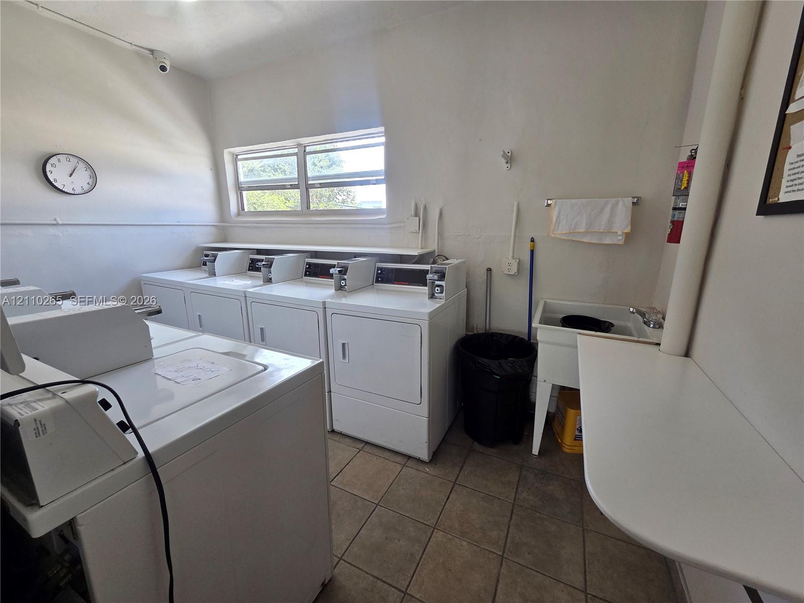 Laundry room featuring tile flooring, multiple washing and drying units, a built-in utility sink, and a wall-mounted folding counter