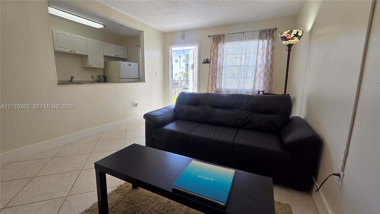 Open-concept interior featuring neutral wall tones, ceramic tile flooring set on the diagonal, and a kitchen pass-through window with built-in cabinetry