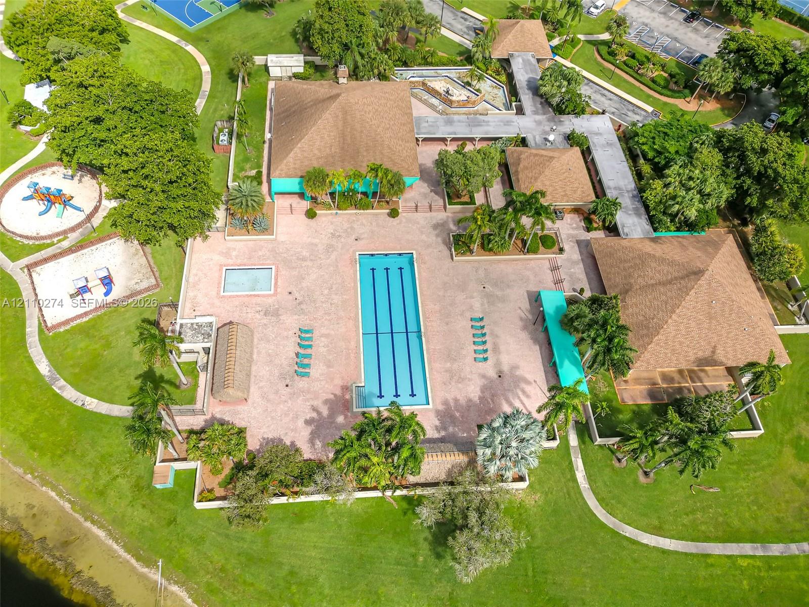 Aerial perspective of a community facility featuring a lap pool, secondary wading pool, paved sundeck, covered walkways, and manicured grounds